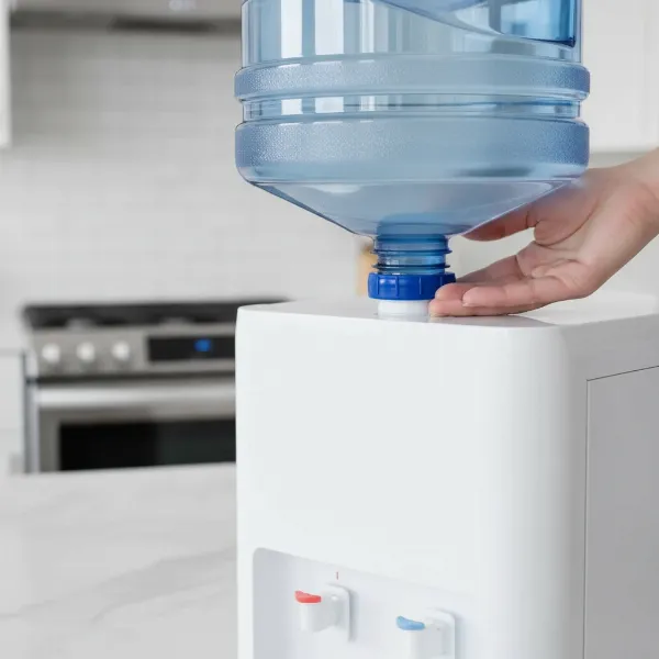 A close-up of a clear 5-gallon water jug with a blue silicone non-spill cap being loaded onto a water dispenser, showing no drips.